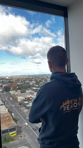 Andy in a North Star Removals hoodie looking out a high-rise apartment window over Melbourne